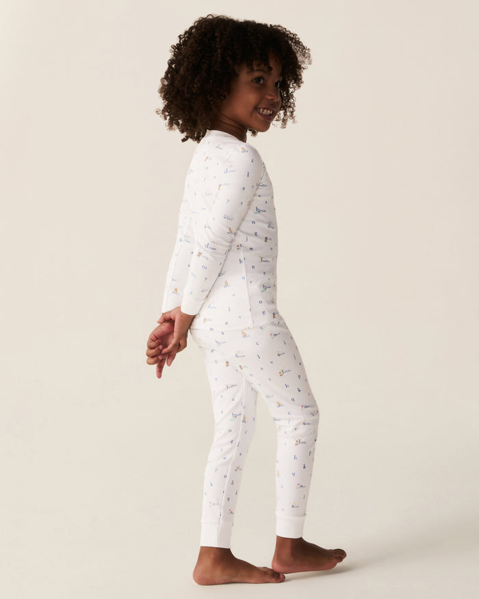 A young child with curly hair stands barefoot in the Kid’s Pima Snug Fit Pajama Set in Letters and Lullabies, smiling and looking to the side with hands clasped behind their back against a plain background.