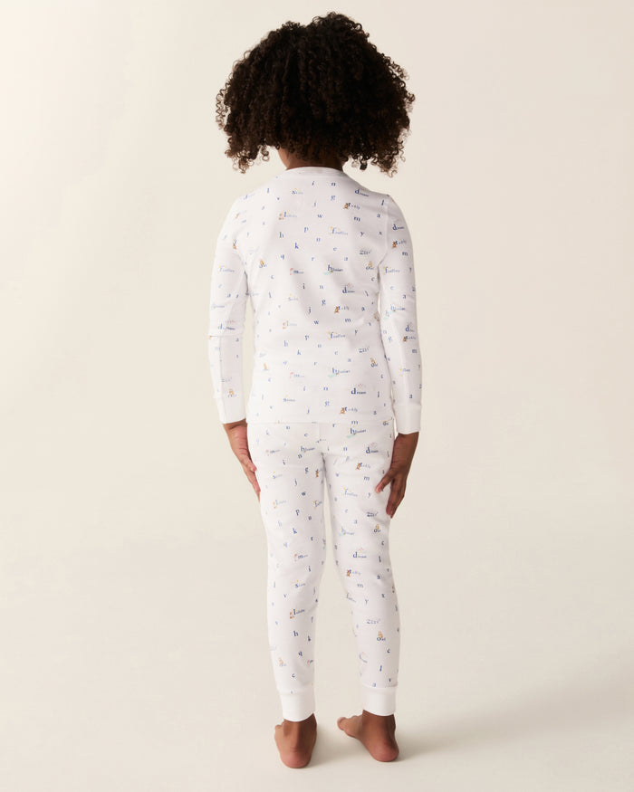 A child with curly hair stands barefoot, facing away, wearing the Kid’s Pima Snug Fit Pajama Set in Letters and Lullabies—white long-sleeve pajamas adorned with small, colorful patterns—against a plain light background.