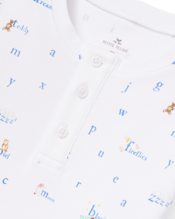 The Kid’s Pima Snug Fit Pajama Set in Letters and Lullabies features a white shirt adorned with blue alphabet letters and small whimsical illustrations, including teddy bears, owls, and fireflies. The design is completed with a three-button placket at the collar.