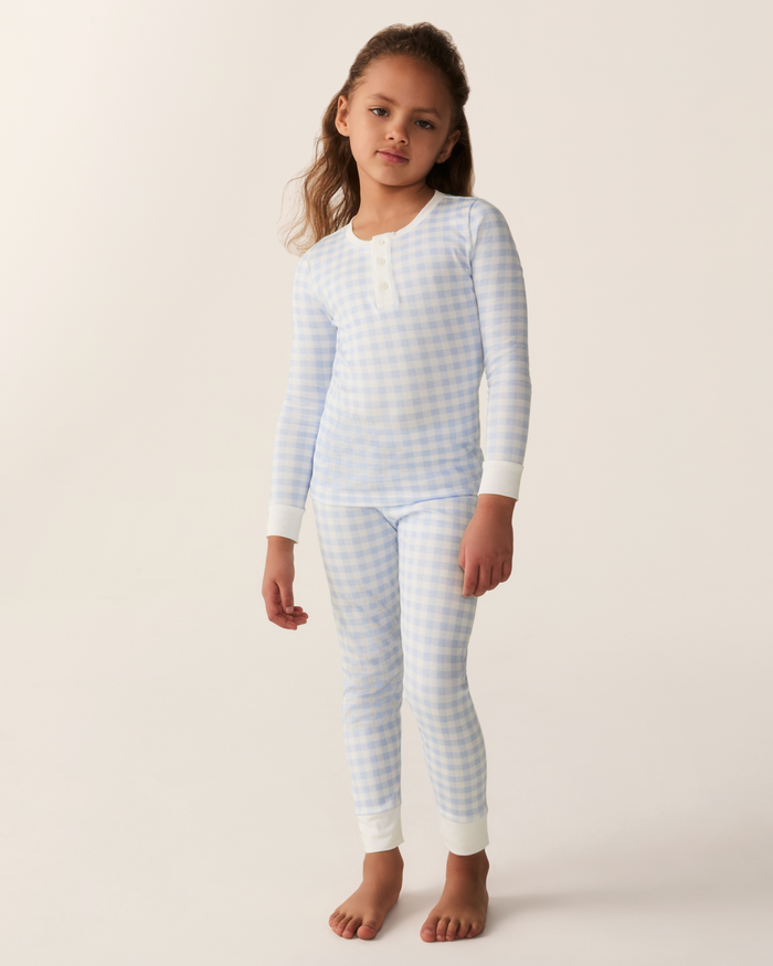 A young child stands barefoot against a plain light background, wearing the Kid’s Pima Snug Fit Pajama Set in Light Blue Gingham—long-sleeve pajamas featuring a light blue and white checkered pattern with white cuffs.