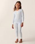 A young child stands barefoot against a plain light background, wearing the Kid’s Pima Snug Fit Pajama Set in Light Blue Gingham—long-sleeve pajamas featuring a light blue and white checkered pattern with white cuffs.