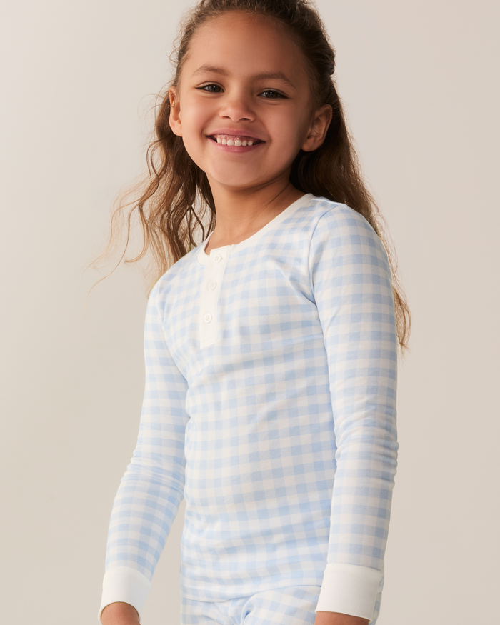 Smiling young girl with long wavy hair wearing the Kid’s Pima Snug Fit Pajama Set in Light Blue Gingham, featuring long sleeves, standing against a neutral background.