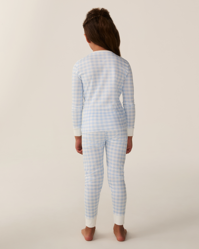 A child stands barefoot facing away, wearing the Kid’s Pima Snug Fit Pajama Set in Light Blue Gingham with long sleeves and long pants, against a plain light background.