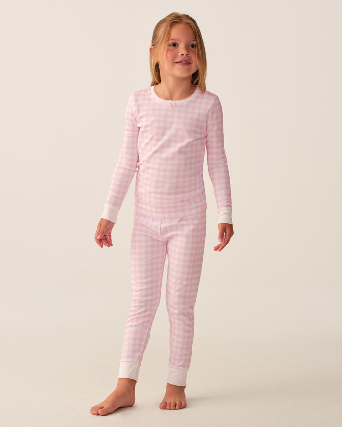 A young girl stands barefoot against a plain light background, wearing the Kid’s Pima Snug Fit Pajama Set in Pink Gingham—featuring pink and white checkered long sleeves with white cuffs.