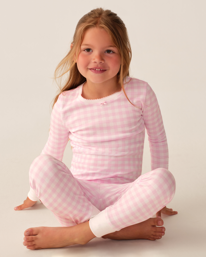 A young girl with long light brown hair sits cross-legged, smiling, and wearing the Kid’s Pima Snug Fit Pajama Set in Pink Gingham, which features a long-sleeve top and matching pants with white cuffs, against a plain light background.