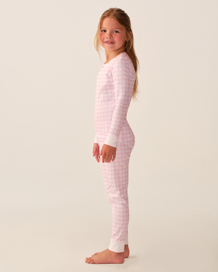 A young girl stands sideways, smiling and barefoot, wearing the Kid’s Pima Snug Fit Pajama Set in Pink Gingham—a long-sleeve pink-and-white checkered set with white cuffs—against a plain light background.