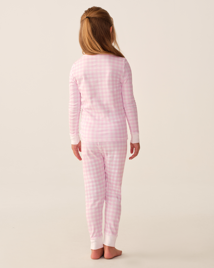 A young girl with light brown hair stands barefoot, facing away, wearing the Kid’s Pima Snug Fit Pajama Set in Pink Gingham against a plain light background.