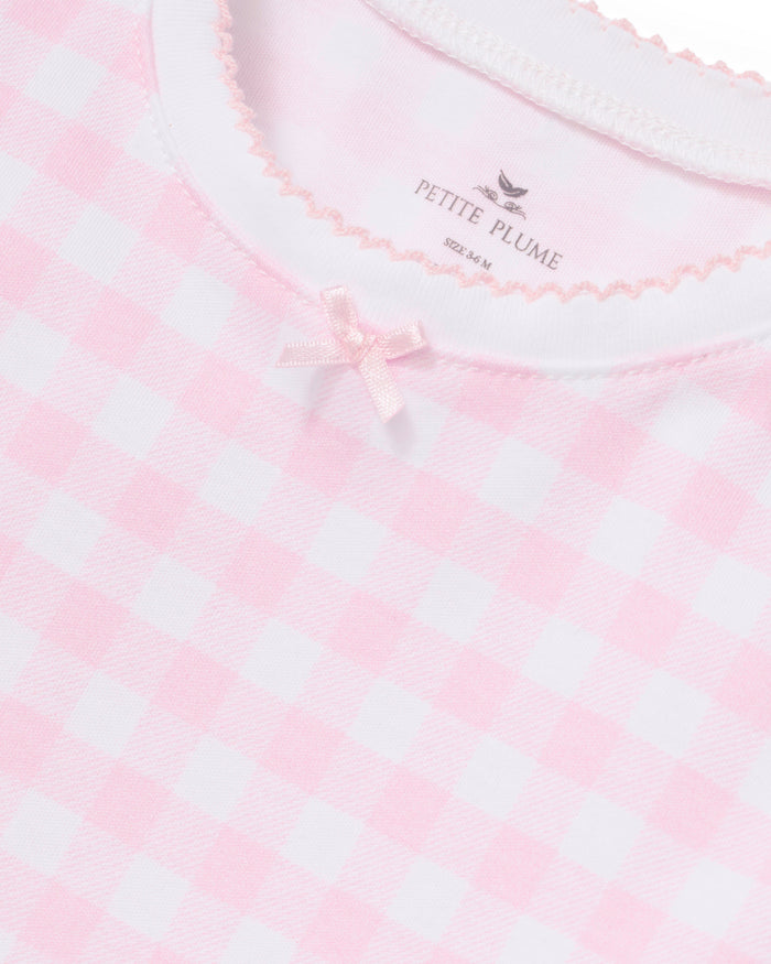 Close-up of the Kid’s Pima Snug Fit Pajama Set in Pink Gingham, featuring a pink and white gingham pattern, a small pink bow at the neckline, and scalloped trim. The inside tag reads Petite Plume.