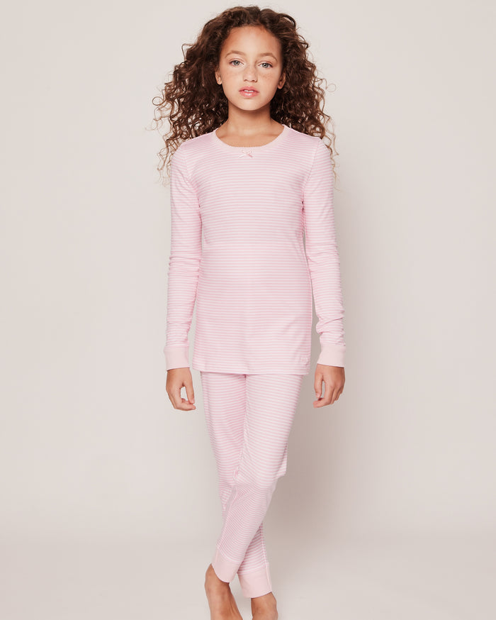 A young girl with long curly hair stands against a plain background, wearing Petite Plumes Kids Pima Snug Fit Pajama Set in Pink Stripes, featuring soft pima cotton and a cozy long-sleeve top with pants.