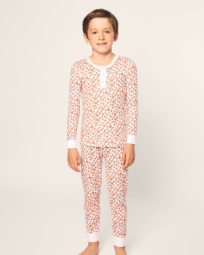 A child models Petite Plumes Kids Pima Snug Fit Pajama Set in Pumpkin Patch. Standing barefoot against a light-colored background, they smile at the camera in cozy, soft Pima Cotton pajamas featuring a playful pumpkin print with a buttoned-up, long-sleeve top.