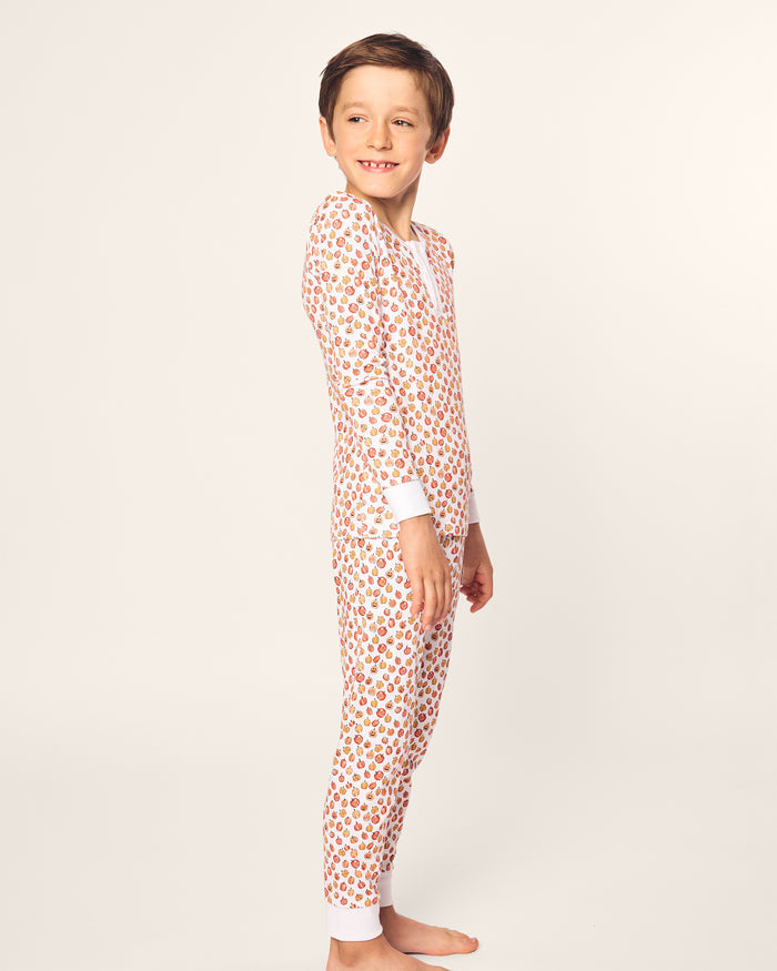 A child with short brown hair smiles while standing sideways, wearing Petite Plumes Kids Pima Snug Fit Pajama Set in Pumpkin Patch. The pajamas feature small red and brown designs on a light background, adding to the cozy vibe as the child stands barefoot against an off-white backdrop.