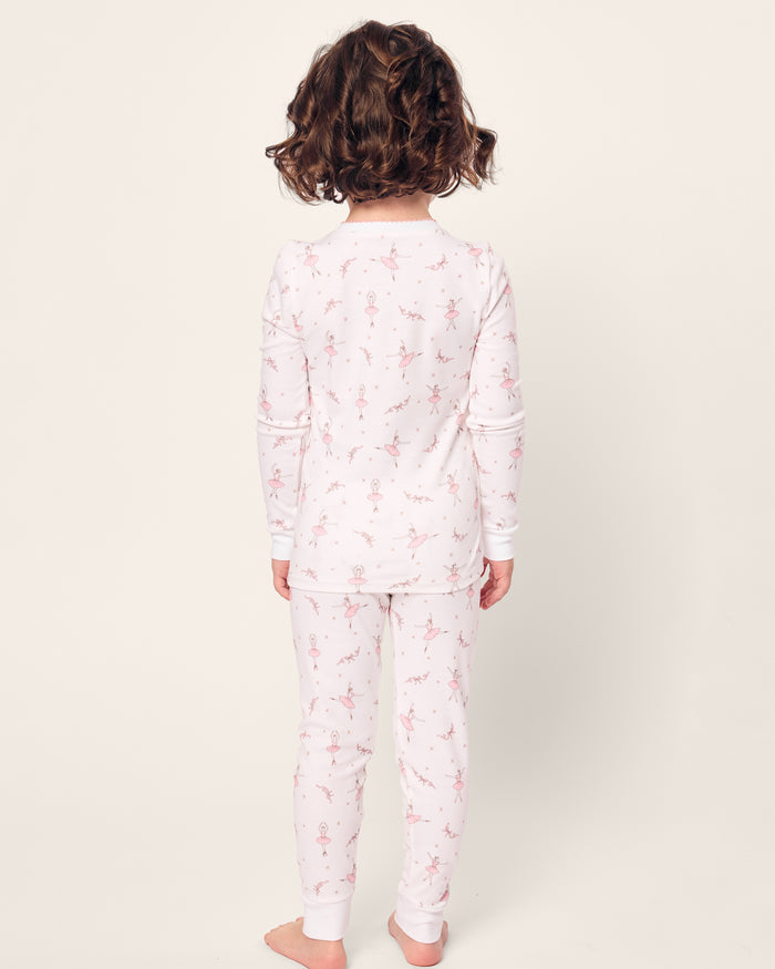 A child with curly hair wears Petite Plumes Kids Pima Snug Fit Pajama Set in Sugar Plum Fairy, featuring soft white Pima cotton and small pink floral designs. They stand barefoot in a long sleeve top, facing away on a plain white background.