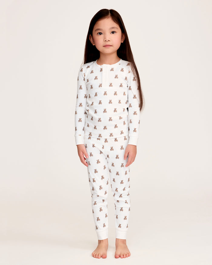 The Kid's Pima Snug Fit Pajama Set in Teddy Tales features a white long-sleeve buttoned top and elastic waist pants with ribbed cuffs, both printed all over with brown teddy bears. Suitable for children.