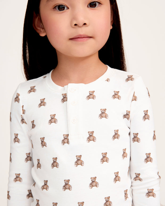 The Kid's Pima Snug Fit Pajama Set in Teddy Tales features a white long-sleeve buttoned top and elastic waist pants with ribbed cuffs, both printed all over with brown teddy bears. Suitable for children.