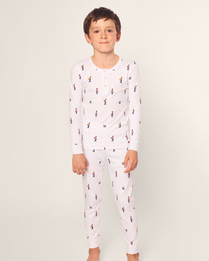 A boy stands barefoot against a plain background, wearing Petite Plumes luxurious Kids Pima Snug Fit Pajama Set in Toy Soldier, featuring small multicolored printed designs.