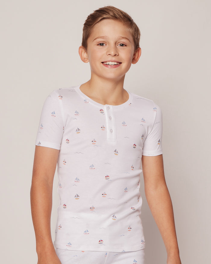 A smiling young boy stands against a plain background, wearing Petite Plumes Kids Pima Snug Fit Pajama Short Set in Bateau, featuring small sailboats. Crafted from soft Pima Cotton, the ensemble includes a buttoned neckline and short sleeves.