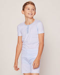 A child in Petite Plumes Kids Pima Snug Fit Pajama Short Set in Blue Stripes smiles and looks aside. The short-sleeve top, crafted from soft Pima cotton, has buttons. The background is plain and light-colored.