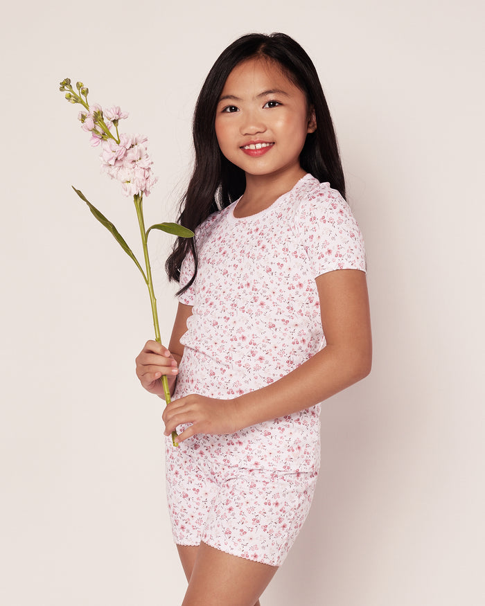 A young girl smiles against a light background in the Kids Pima Snug Fit Pajama Short Set in Dorset Floral by Petite Plume, holding a light pink flower with green leaves. Her short sleeve top beautifully complements the gentle hues around her.