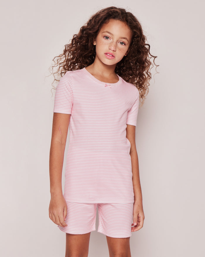 A young girl with curly hair wears the Petite Plume Kids Pima Snug Fit Pajama Short Set in Pink Stripes, gazing neutrally at the camera against a light background, highlighting her adorable sleepwear look.