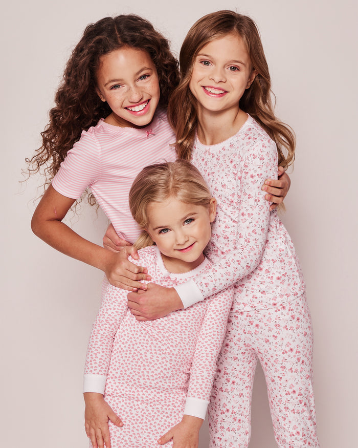 Three young girls in Petite Plume coordinating Kids Pima Snug Fit Pajama Short Sets. The two older, long-haired girls wear pink stripes, and the younger matches in a similar style. All are smiling and hugging against a plain backdrop.