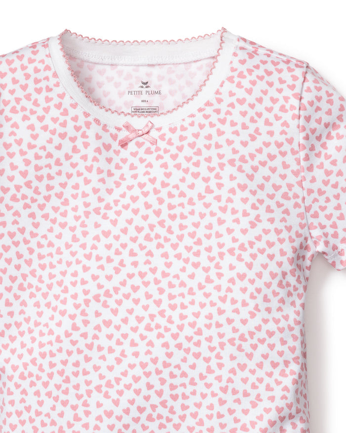 The Kids Pima Snug Fit Pajama Short Set in Sweethearts from Petite Plume features a white T-shirt with a pink heart pattern, a pink bow at the neckline, and scalloped trim. Made from soft Pima Cotton Sleepwear, its ideal for childrens luxury sleepwear. The fabric label inside shows brand details.