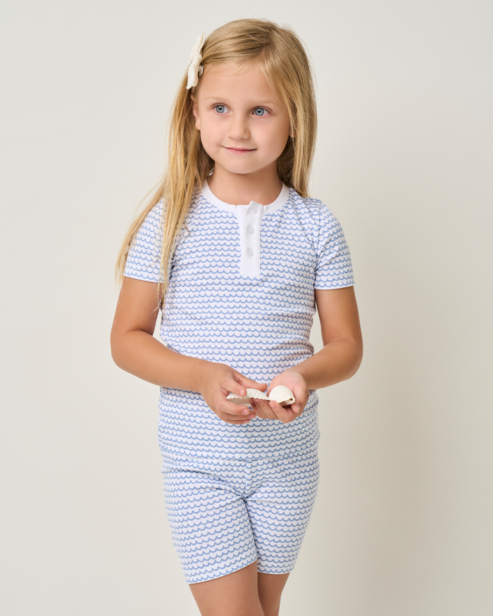 A young girl with long blonde hair stands against a plain background, wearing Petite Plumes Kids Pima Snug Fit Pajama Short Set in La Mer, featuring a small wave pattern. She holds seashells and has a white bow in her hair.