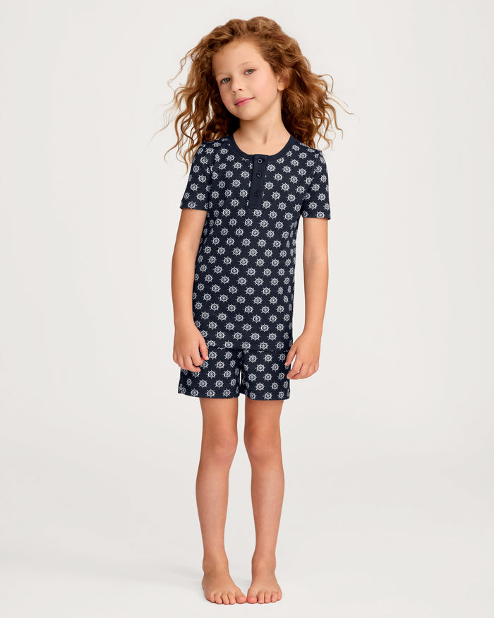 A young child with curly hair stands barefoot in front of a plain light background, wearing the Kid’s Pima Snug Fit Short Set in Captain’s Compass.
