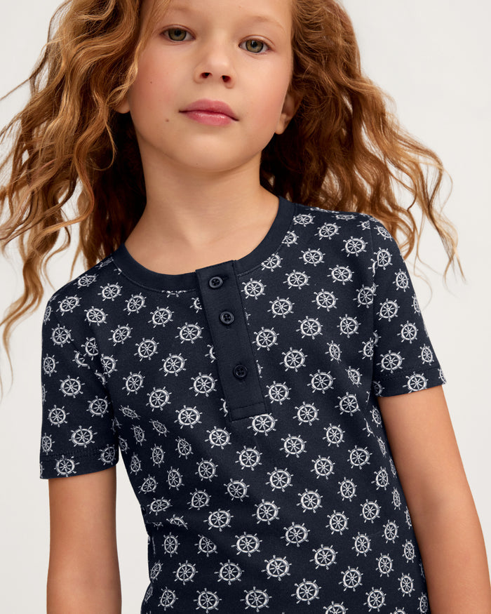 Wearing the Kid’s Pima Snug Fit Short Set in Captain’s Compass, a child with long, wavy hair stands against a light background, showcasing the navy shirt adorned with a ship wheel pattern and three buttons at the collar.