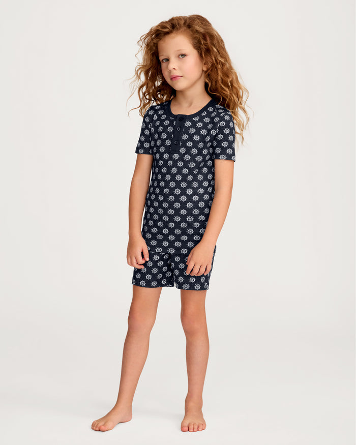 A young girl with curly hair stands barefoot against a plain light background, wearing the Kid’s Pima Snug Fit Short Set in Captain’s Compass, featuring a dark blue short-sleeved top and matching shorts with a white floral pattern.