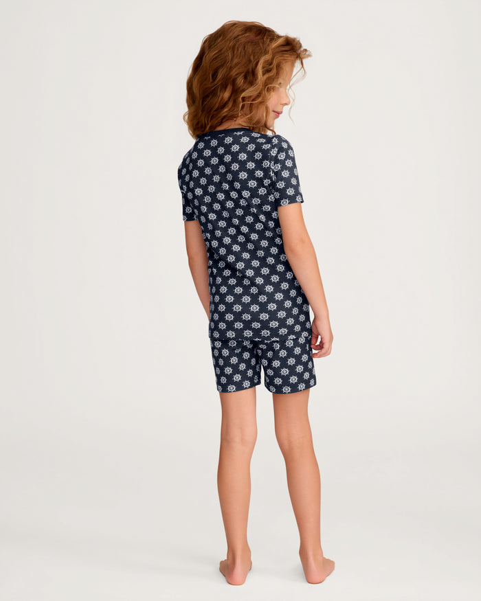 A child with wavy hair stands barefoot, facing away, wearing the Kid’s Pima Snug Fit Short Set in Captain’s Compass—a navy blue short set featuring a white anchor pattern—against a plain light background.
