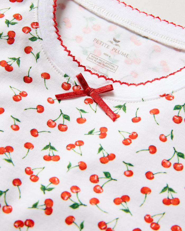 A close-up of the Kids Pima Snug Fit Short Set in Cherry Orchard by Petite Plume shows a white fabric with small red cherries, a red ribbon at the neck, and red-stitched collar. A partially visible label displays branding inside.