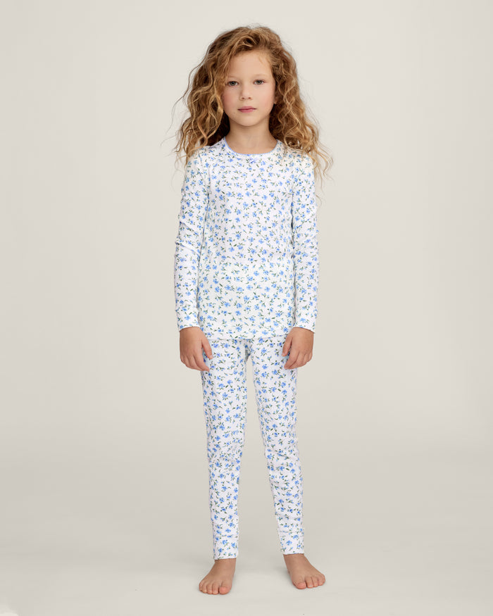 A young child with long curly hair stands barefoot against a plain background, wearing the Kid's Pointelle Snug Fit Pajama Set in Blue Floral.