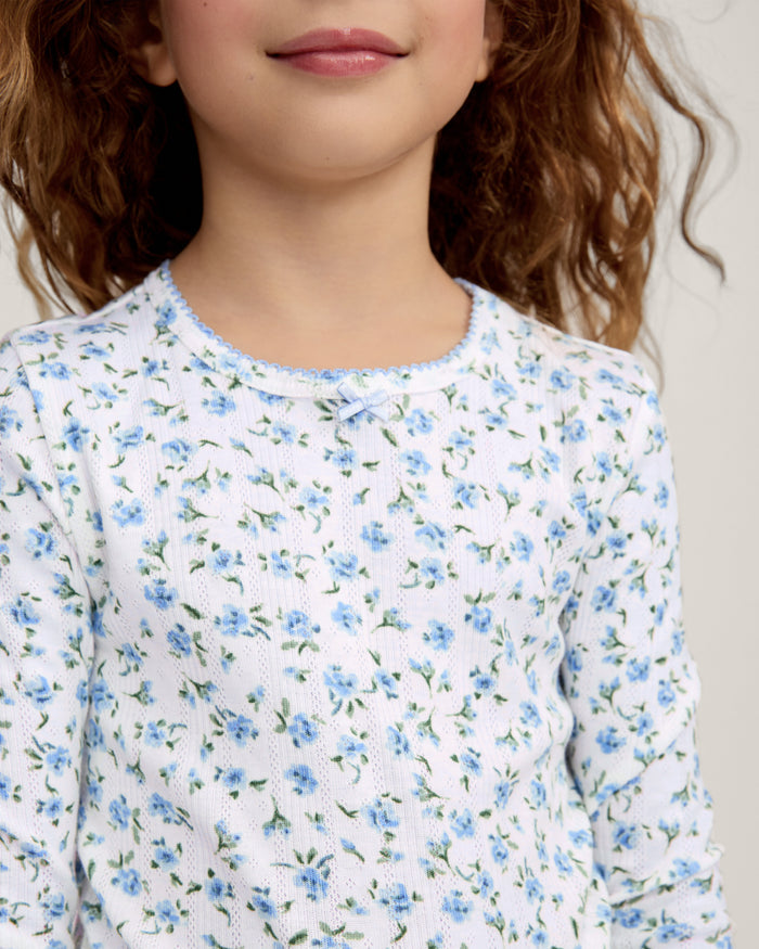 A young child with long curly hair stands barefoot against a plain background, wearing the Kid's Pointelle Snug Fit Pajama Set in Blue Floral.