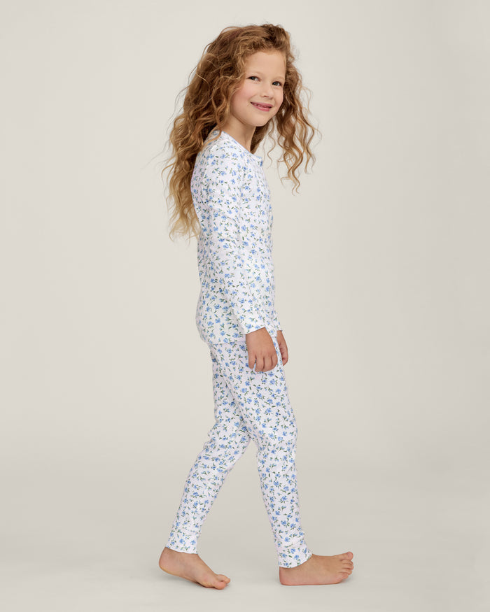A young child with long curly hair stands barefoot against a plain background, wearing the Kid's Pointelle Snug Fit Pajama Set in Blue Floral.