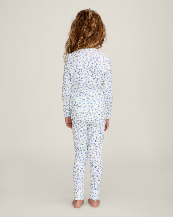 A young child with long curly hair stands barefoot against a plain background, wearing the Kid's Pointelle Snug Fit Pajama Set in Blue Floral.