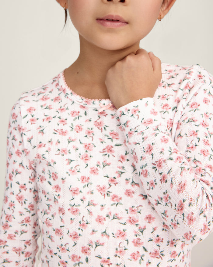 A young girl stands barefoot against a plain background, wearing the Kid's Pointelle Snug Fit Pajama Set in Pink Floral. She faces forward with a relaxed, neutral expression.