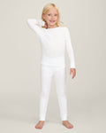 A happy child with long blonde hair stands barefoot against a plain background, wearing the Kid's Pointelle Snug Fit Pajama Set in White—long-sleeve top and matching pants—with one hand behind their head and the other arm relaxed.