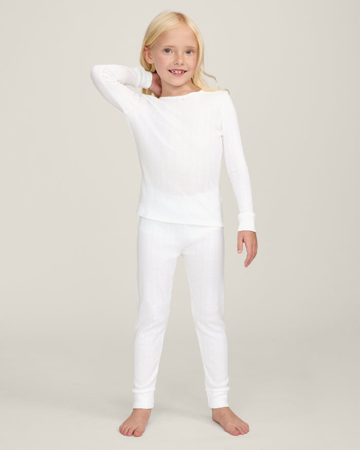A happy child with long blonde hair stands barefoot against a plain background, wearing the Kid's Pointelle Snug Fit Pajama Set in White—long-sleeve top and matching pants—with one hand behind their head and the other arm relaxed.
