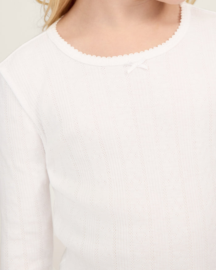 A happy child with long blonde hair stands barefoot against a plain background, wearing the Kid's Pointelle Snug Fit Pajama Set in White—long-sleeve top and matching pants—with one hand behind their head and the other arm relaxed.