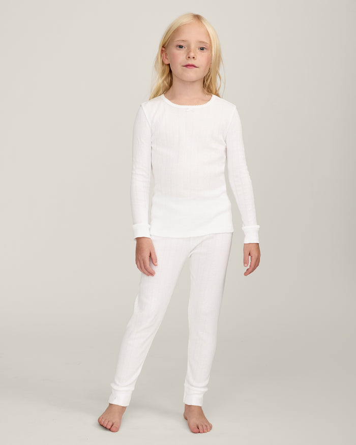A happy child with long blonde hair stands barefoot against a plain background, wearing the Kid's Pointelle Snug Fit Pajama Set in White—long-sleeve top and matching pants—with one hand behind their head and the other arm relaxed.