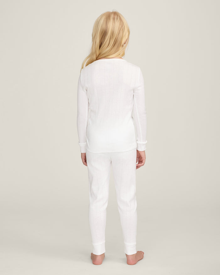 A happy child with long blonde hair stands barefoot against a plain background, wearing the Kid's Pointelle Snug Fit Pajama Set in White—long-sleeve top and matching pants—with one hand behind their head and the other arm relaxed.