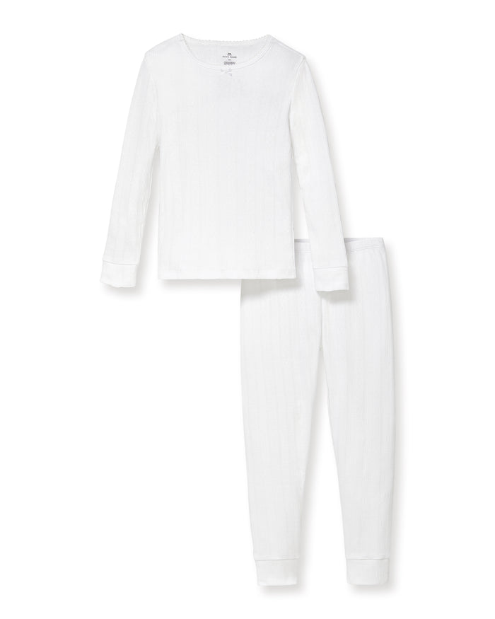 A happy child with long blonde hair stands barefoot against a plain background, wearing the Kid's Pointelle Snug Fit Pajama Set in White—long-sleeve top and matching pants—with one hand behind their head and the other arm relaxed.