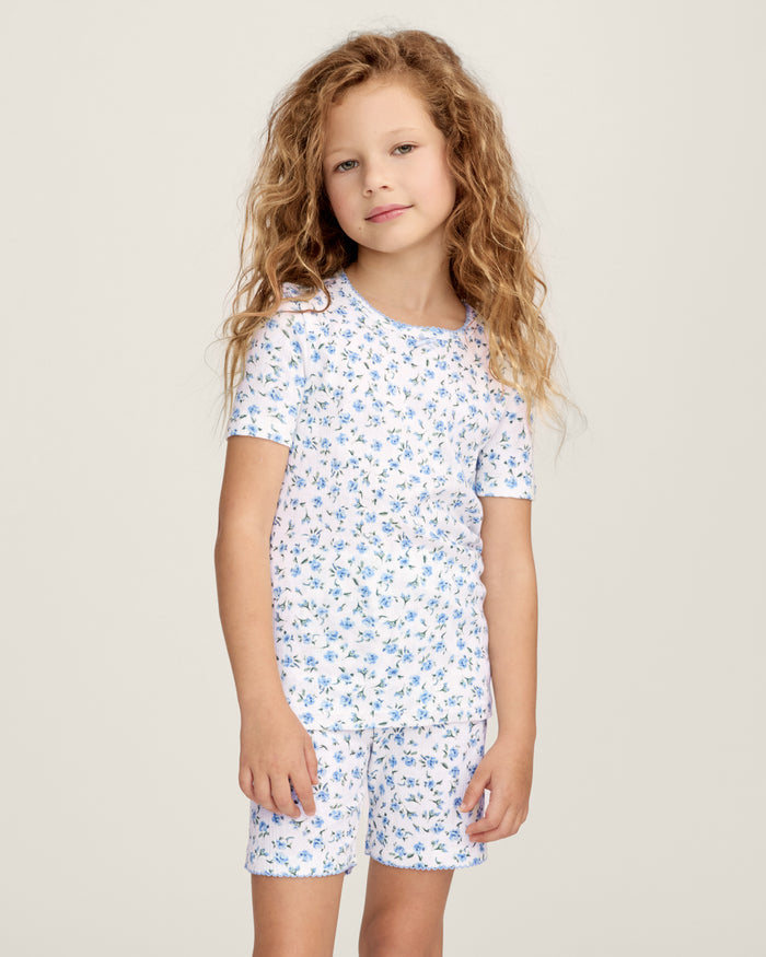 A young girl with long, curly light brown hair stands against a plain background, wearing the Kid's Pointelle Snug Fit Pajama Short Set in Blue Floral. She looks at the camera with a gentle expression.