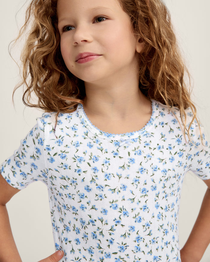 A young girl with long, curly light brown hair stands against a plain background, wearing the Kid's Pointelle Snug Fit Pajama Short Set in Blue Floral. She looks at the camera with a gentle expression.
