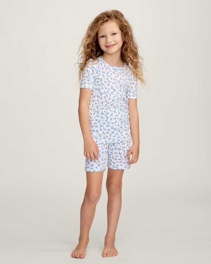 A young girl with long, curly light brown hair stands against a plain background, wearing the Kid's Pointelle Snug Fit Pajama Short Set in Blue Floral. She looks at the camera with a gentle expression.