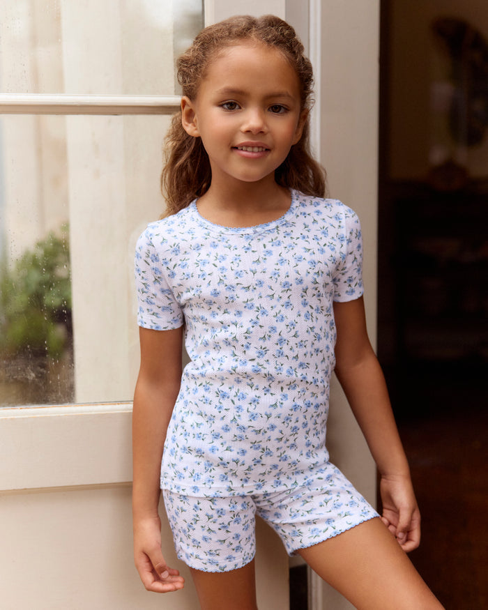 A young girl with long, curly light brown hair stands against a plain background, wearing the Kid's Pointelle Snug Fit Pajama Short Set in Blue Floral. She looks at the camera with a gentle expression.