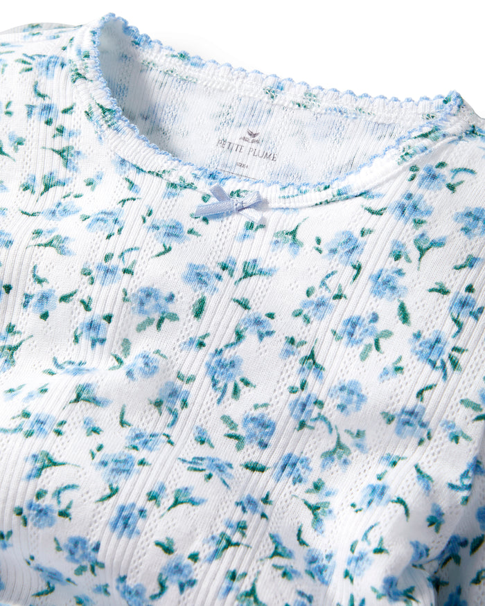 A young girl with long, curly light brown hair stands against a plain background, wearing the Kid's Pointelle Snug Fit Pajama Short Set in Blue Floral. She looks at the camera with a gentle expression.
