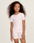 A young girl with curly hair stands against a plain background, wearing the Kid's Pointelle Snug Fit Pajama Short Set in Pink Floral, featuring a matching short-sleeve top and shorts decorated with a pink and red floral pattern.