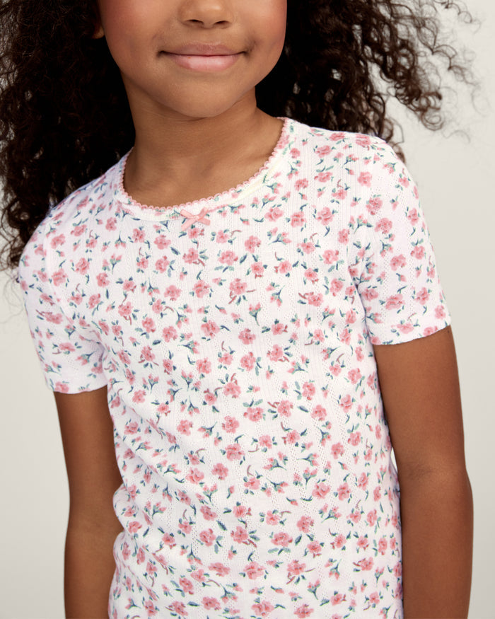 A young girl with curly hair stands against a plain background, wearing the Kid's Pointelle Snug Fit Pajama Short Set in Pink Floral, featuring a matching short-sleeve top and shorts decorated with a pink and red floral pattern.