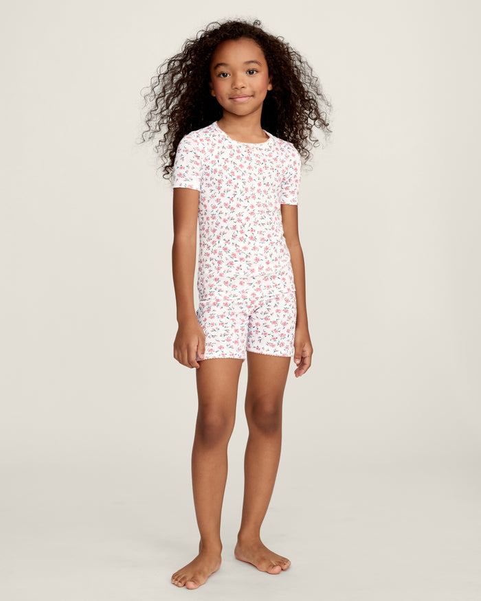 A young girl with curly hair stands against a plain background, wearing the Kid's Pointelle Snug Fit Pajama Short Set in Pink Floral, featuring a matching short-sleeve top and shorts decorated with a pink and red floral pattern.